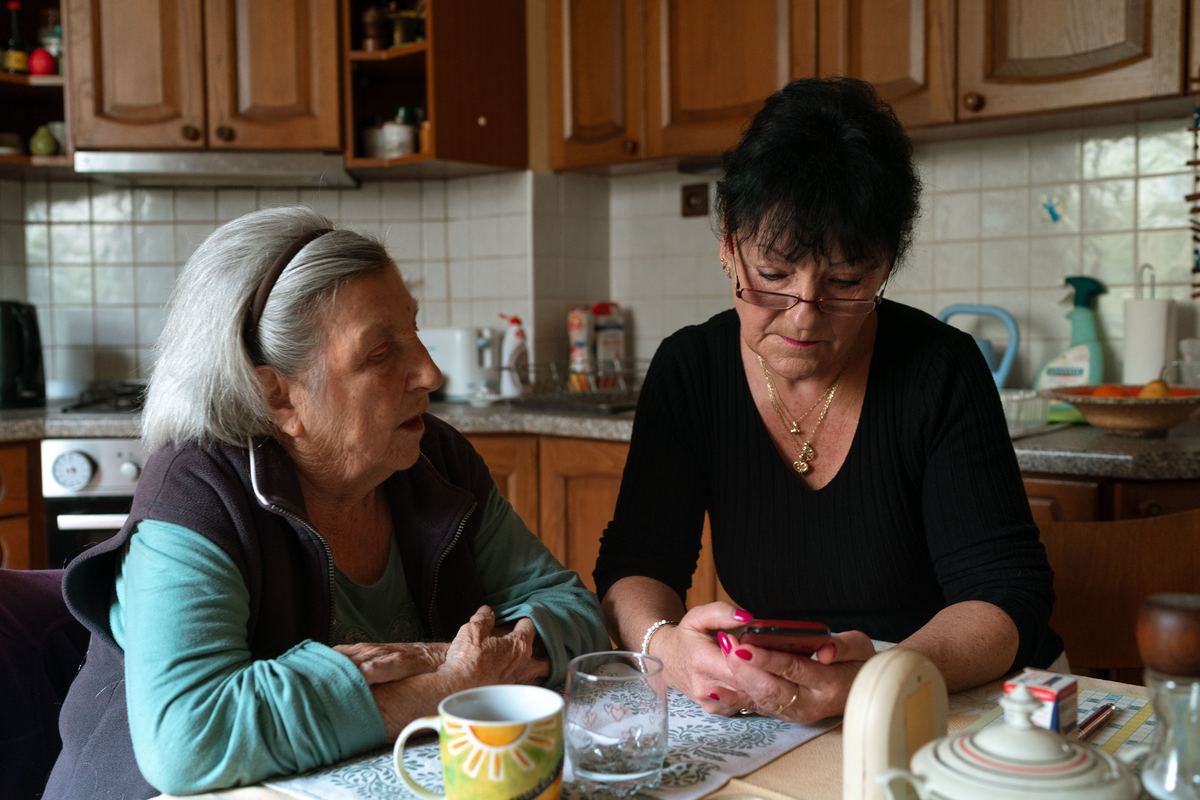 Een oudere vrouw en een zorgverleenster praten aan de keukentafel.